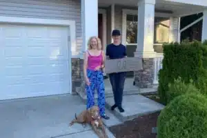 Two people holding a ‘Sold’ sign outside a home, with a dog beside them